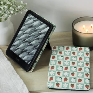 May include: A Kindle e-reader in a protective case featuring a light green and white checkered pattern with red strawberries and white flowers. One device is propped open displaying a grayscale image, while another is closed showing the same pattern.