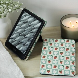 May include: A Kindle e-reader in a protective case featuring a light green and white checkered pattern with red strawberries and white flowers. One device is propped open displaying a grayscale image, while another is closed showing the same pattern.