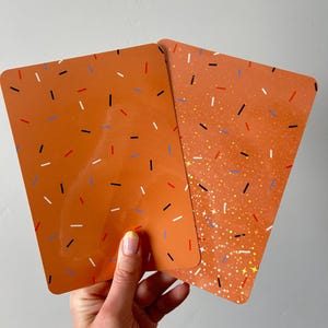 Op de afbeelding: Twee rechthoekige kaarten met afgeronde hoeken, een oranje achtergrond en een patroon van kleine, verspreide streepjes in zwart, wit, rood en blauw. De ene kaart heeft een gladde matte afwerking, terwijl de andere een holografische glitterlaag heeft.