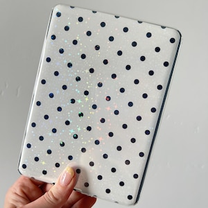 Puede incluir: Una funda protectora rectangular para lector electrónico o tableta con fondo blanco, un patrón repetitivo de pequeños puntos negros y un sutil efecto de estrellas holográficas que capta la luz sobre la superficie de la cubierta.