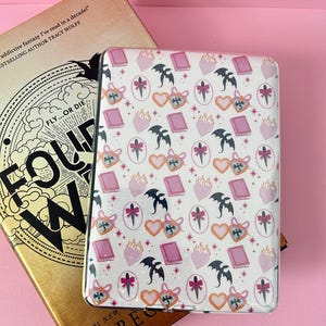 Könnte beinhalten: Eine rechteckige E-Reader-Hülle mit weißem Hintergrund und einem wiederkehrenden Muster aus rosa Herzen, schwarzen Drachensilhouetten, rosa Büchern und Dolchen mit rosa Schleifen. Die Hülle liegt auf einem goldenen Buchumschlag mit dem Text FOUR WING.