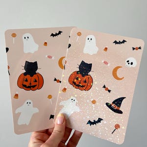 Op de afbeelding: Twee rechthoekige stickers met een lichtroze achtergrond met een herhalend Halloween-patroon van witte spoken, zwarte katten op oranje pompoenen, vleermuizen, snoepjes, lolly's en een heksenhoed. Eén sticker heeft een holografische glitterafwerking.