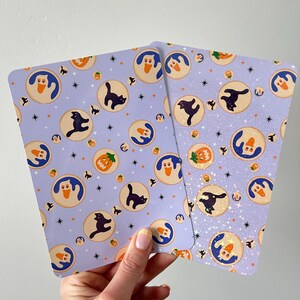 May include: Two rectangular cards with a light purple background featuring a repeating pattern of circular icons containing white ghosts, black cats, and orange jack-o'-lanterns, interspersed with small black stars and sparkles.