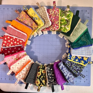 May include: A circular arrangement of twenty-one small, rectangular fabric zipper pouches. Each pouch features a unique pattern, including floral, gingham, polka dot, and solid prints in various colors like red, yellow, blue, green, and purple.