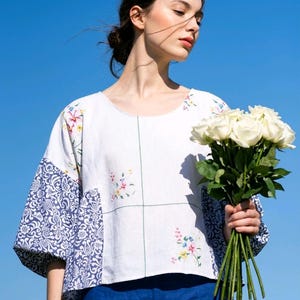 May include: A white blouse featuring a round neckline, three-quarter length sleeves with blue and white floral patterned fabric, and embroidered floral accents on the front and shoulders, paired with solid blue trousers.