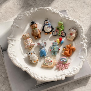 May include: A collection of eleven small, colorful clay charms featuring a bear in a top hat, a penguin with a scarf, a frog on a bicycle, a hedgehog, a cat in a floral dress, a mouse in overalls, a robot with balloons, an owl, a snail, and an octopus.