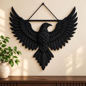 Op de afbeelding: Een zwarte gehaakte wandhanger die een raaf met gespreide vleugels voorstelt. De vogel hangt aan een horizontale zwarte roede met een driehoekige koordhanger aan de bovenkant. De gedetailleerde textuur van de veren is gecreëerd door complexe haaksteken, wat een symmetrisch vogel silhouet vormt.
