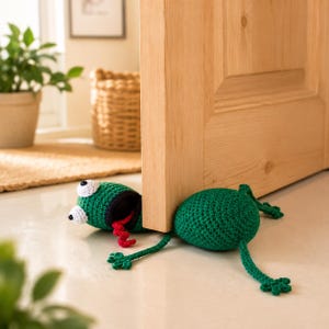 Puede incluir: Un tope de puerta con forma de rana tejida a ganchillo en color verde, colocado bajo una puerta de madera. La rana tiene ojos blancos con pupilas negras, una boca negra y una larga lengua roja tejida que sobresale. Cuerpo redondeado.
