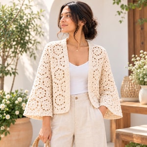 May include: An off-white, open-front crochet cardigan featuring a repeating hexagonal floral pattern, wide three-quarter length sleeves, and a straight hem, worn over a simple white camisole and light-colored high-waisted trousers.