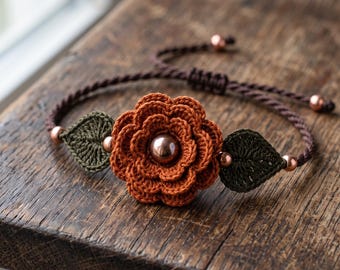 May include: A bracelet featuring a central crocheted orange flower with a metallic copper bead at its center, flanked by two dark green crocheted leaves. The band is made of dark brown twisted cord with small copper beads and an adjustable closure.