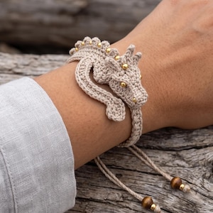 May include: A beige crochet bracelet featuring a three-dimensional dragon design. The dragon has small gold-colored beads along its back and head, with gold-colored bead eyes. The bracelet includes adjustable cords finished with two brown wooden beads and gold-colored accents.
