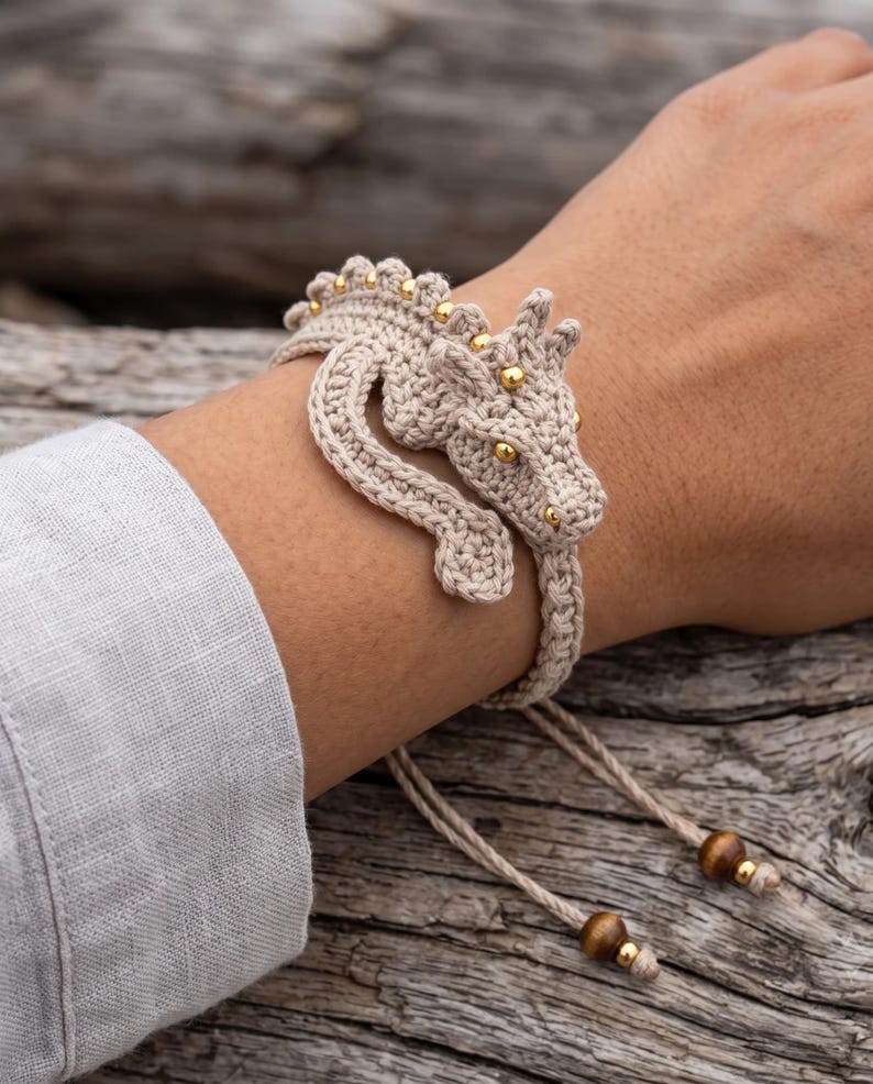 May include: A beige crochet bracelet featuring a three-dimensional dragon design. The dragon has small gold-colored beads along its back and head, with gold-colored bead eyes. The bracelet includes adjustable cords finished with two brown wooden beads and gold-colored accents.