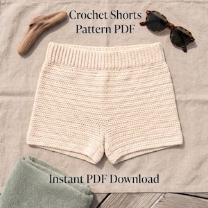 May include: Cream-colored crochet shorts with a ribbed waistband, accompanied by a piece of driftwood and tortoise-shell sunglasses. The text reads Crochet Shorts Pattern PDF and Instant PDF Download.