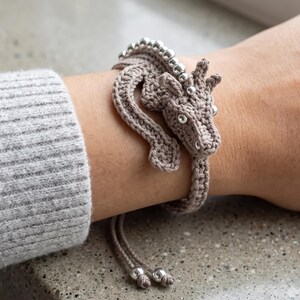 May include: A beige crocheted dragon bracelet featuring a long, winding body that wraps around the wrist. The dragon has a detailed head with small silver beads for eyes and a row of silver beads along its back. Two thin cords with silver beads hang down.