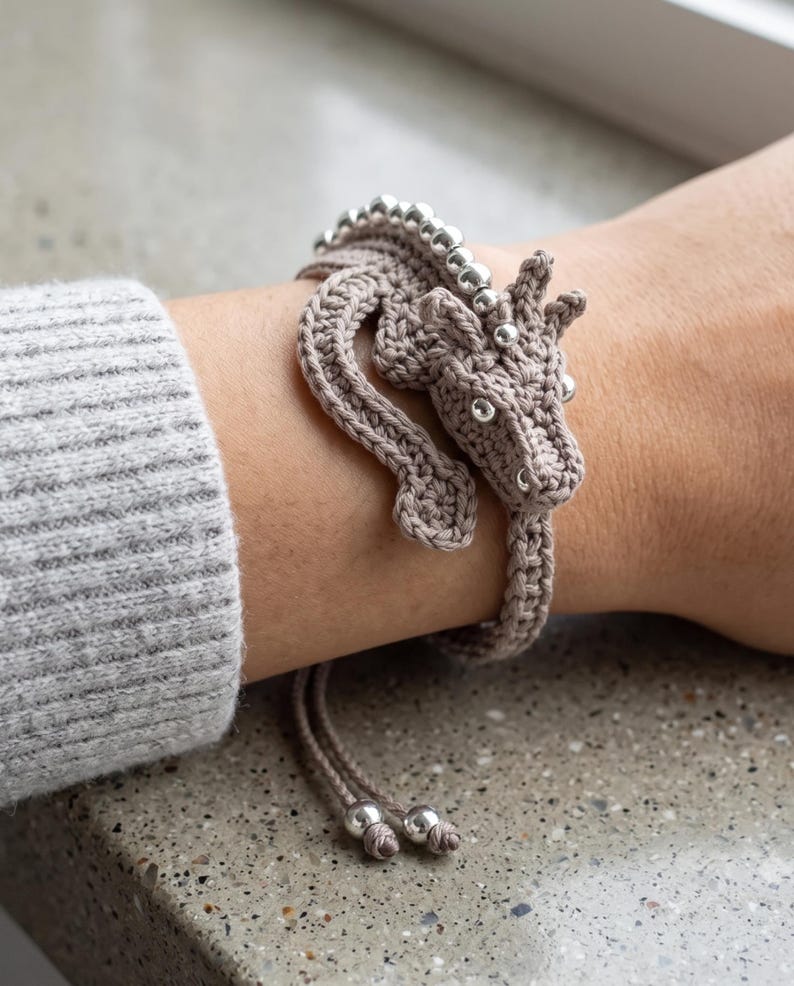 May include: A beige crocheted dragon bracelet featuring a long, winding body that wraps around the wrist. The dragon has a detailed head with small silver beads for eyes and a row of silver beads along its back. Two thin cords with silver beads hang down.