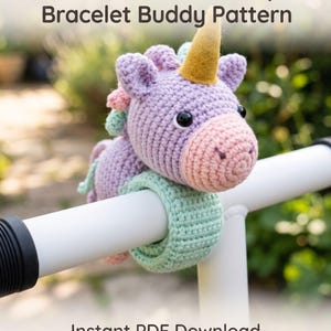 Puede incluir: Una pulsera de bofetada de unicornio de ganchillo con cabeza lavanda, hocico rosa, cuerno dorado y banda verde menta. El texto dice: Crochet Unicorn Slap Bracelet Buddy Pattern e Instant PDF Download. El unicornio tiene ojos negros y crin.