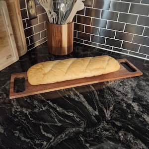 May include: A long, golden-brown loaf of bread with diagonal score marks rests on a rectangular wooden serving board. The board features integrated handles on both ends and a natural wood grain finish, providing a stable base for the bread.