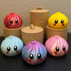 May include: A collection of five colorful, spherical dumpling figurines with smiling faces, large black eyes, and red cheeks. Each dumpling sits atop or beside a tan, cylindrical bamboo-style steamer basket with a woven lid and handle.