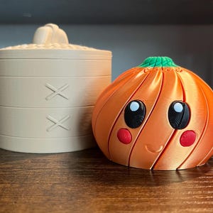 May include: A beige cylindrical container with a lid featuring two X-shaped markings and a textured top, positioned next to a bright orange pumpkin-shaped object with a green stem, black eyes, and small red circular cheeks on its front surface.