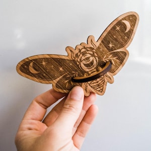 May include: Wooden moth-shaped phone stand with intricate celestial design. The stand features a crescent moon, stars, and a rose. The wings are detailed with etched patterns. The stand is held by a hand.