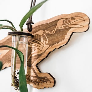 May include: A wooden wall hanging in the shape of a raven with a glass test tube for a plant. The raven is laser-cut and has a detailed skeleton design. The test tube is filled with water and a small plant with green leaves.