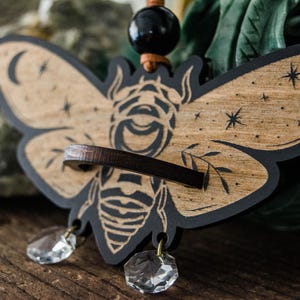 May include: Wooden moth pendant with a black outline and a natural wood grain finish. The moth design features a crescent moon, stars, and a central eye. Two clear crystal pendants hang below. The pendant is suspended from a black bead and leather cord.