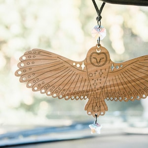 May include: Wooden owl car charm with etched details. The owl has outstretched wings and star accents. A faceted crystal hangs below the owl and another above. The charm is suspended from a black cord, designed to hang from a car's rearview mirror.
