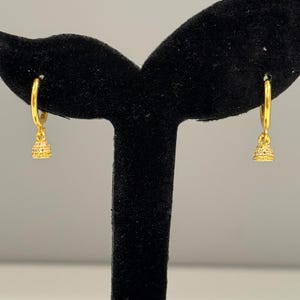 May include: A pair of gold hoop earrings featuring small, bell-shaped charms. Each charm is textured with a fine, intricate pattern and hangs from a polished gold hoop. The earrings are displayed on a black stand, highlighting the metallic finish.