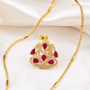 May include: A gold-toned necklace featuring a pendant shaped like the deity Ganesha, adorned with multiple pear-cut red stones and small clear pavé crystals. The chain is a delicate gold-toned link style with smooth cylindrical accents throughout.