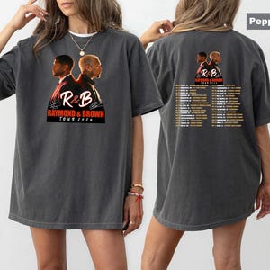 May include: A charcoal gray oversized t-shirt featuring a graphic of two men back-to-back with the text R&B RAYMOND & BROWN TOUR 2026 on the front. The back displays the same graphic with a list of tour dates and locations printed in white text.