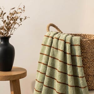 Op de afbeelding: Een zachte, saliegroene handdoek met dunne, horizontale bruine strepen. De handdoek is over de rand van een gevlochten rieten mand gedrapeerd en toont een getextureerd, pluche stoffen oppervlak met een consistent patroon van smalle lijnen.