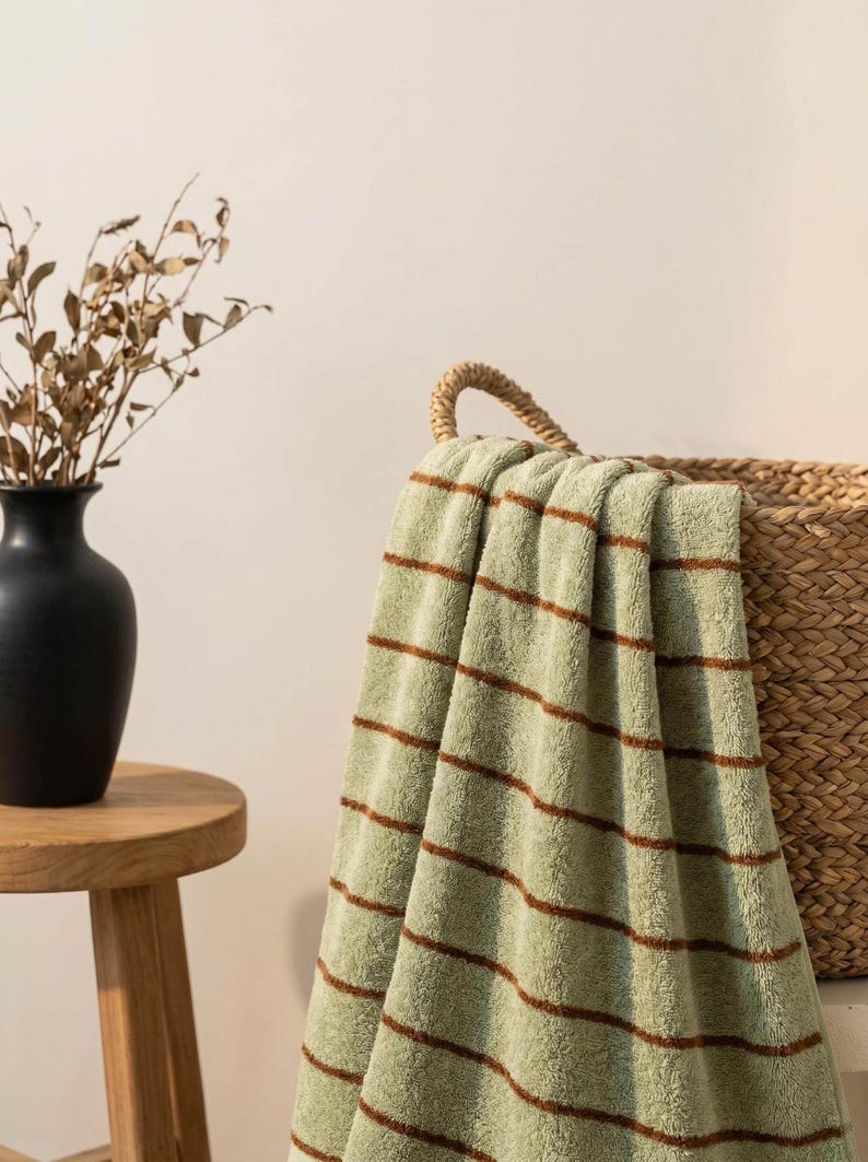 Op de afbeelding: Een zachte, saliegroene handdoek met dunne, horizontale bruine strepen. De handdoek is over de rand van een gevlochten rieten mand gedrapeerd en toont een getextureerd, pluche stoffen oppervlak met een consistent patroon van smalle lijnen.