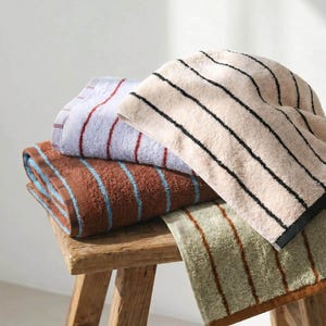 Op de afbeelding: Een stapel van vier gevouwen katoenen handdoeken met diverse streeppatronen. De handdoeken omvatten een lichtblauwe handdoek met rode strepen, een bruine met blauwe strepen, een beige met zwarte strepen en een saliegroene met bruine strepen.