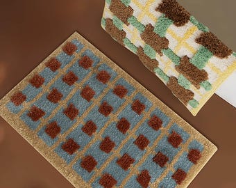 Op de afbeelding: Twee rechthoekige badmatten met een getuft ruitpatroon. De ene mat heeft een blauw en bruin ruitontwerp, terwijl de andere een groen, geel en bruin ruitpatroon vertoont. Beide matten hebben een getextureerd, pluizig oppervlak.