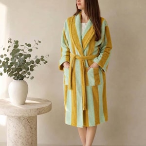 May include: A knee-length towelling bathrobe featuring vertical stripes in mustard yellow and light mint green. The robe includes a wide shawl collar, long sleeves, two front patch pockets, and a matching fabric tie belt at the waist.