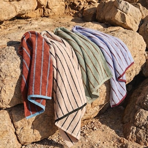Op de afbeelding: Vier rechthoekige katoenen handdoeken met verticale strepen in roestoranje met lichtblauw, beige met zwart, saliegroen met donkergroen en lichtblauw met bordeauxrood. Elke handdoek heeft een contrasterende zoom aan de onderkant.