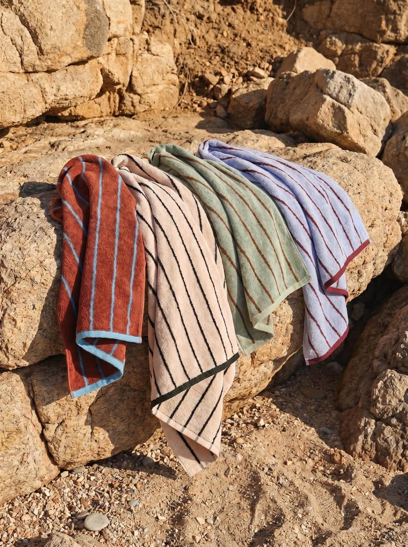 Op de afbeelding: Vier rechthoekige katoenen handdoeken met verticale strepen in roestoranje met lichtblauw, beige met zwart, saliegroen met donkergroen en lichtblauw met bordeauxrood. Elke handdoek heeft een contrasterende zoom aan de onderkant.