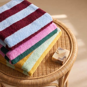 May include: A stack of three folded rectangular towels featuring bold horizontal stripes in alternating colors of light blue, burgundy, pink, dark green, light green, and mustard yellow, accompanied by a single rectangular bar of marbled soap.