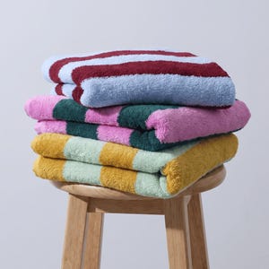 May include: A stack of three folded cotton towels with thick vertical stripes. The top towel features maroon and light blue stripes, the middle towel has dark green and pink stripes, and the bottom towel displays mustard yellow and light green stripes.
