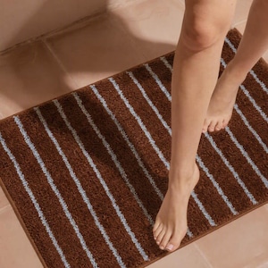 May include: A rectangular bath mat featuring a deep brown, textured pile surface accented with thin, vertical light blue stripes. The mat has a finished edge border in a matching brown tone, providing a clean and structured look for the floor.