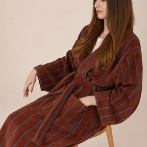 May include: A long-sleeved, knee-length dressing gown in a deep rust-brown colour featuring vertical teal stripes. The robe includes a matching tie belt at the waist, wide sleeves, and two front patch pockets, crafted from a textured, plush fabric.