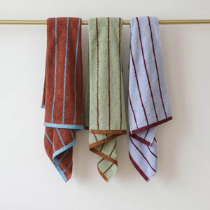 Op de afbeelding: Drie rechthoekige katoenen handdoeken hangen aan een goudkleurige metalen stang. De eerste handdoek is roestkleurig met lichtblauwe verticale strepen, de tweede is saliegroen met bruine strepen en de derde is lichtblauw met donkerrode verticale strepen.