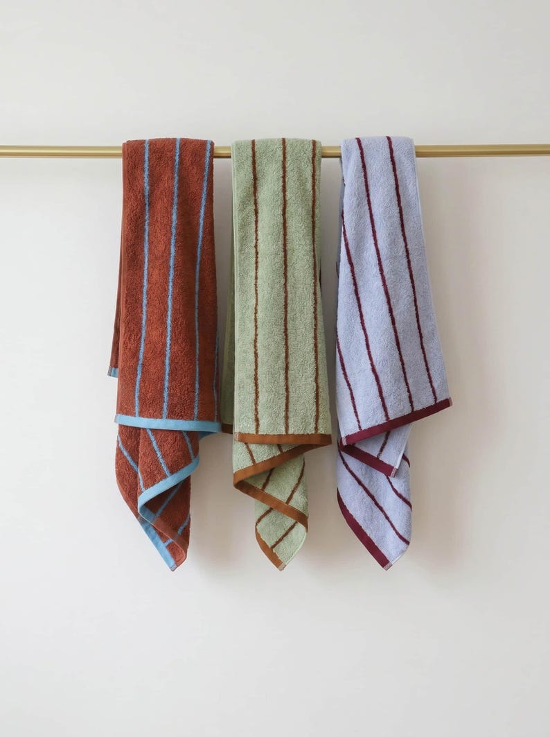 Op de afbeelding: Drie rechthoekige katoenen handdoeken hangen aan een goudkleurige metalen stang. De eerste handdoek is roestkleurig met lichtblauwe verticale strepen, de tweede is saliegroen met bruine strepen en de derde is lichtblauw met donkerrode verticale strepen.
