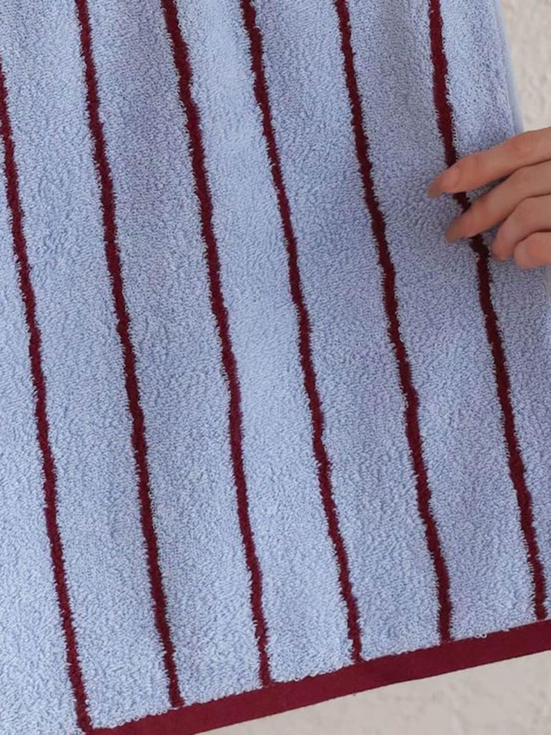 Op de afbeelding: Een lichtblauwe handdoek met verticale donkerrode strepen en een effen donkerrode rand langs de onderkant. De stof heeft een getextureerd oppervlak met lussen en het ontwerp is consistent over het gehele zichtbare deel van het textiel.
