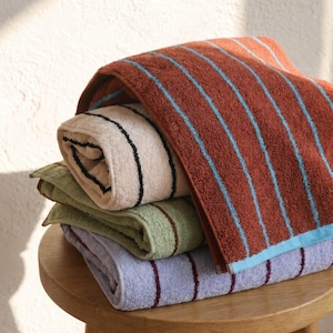 Op de afbeelding: Een stapel van vier gevouwen katoenen handdoeken met verticale strepen. De bovenste handdoek is roestoranje met lichtblauwe strepen, gevolgd door een cr&egrave;mekleurige handdoek met zwarte strepen, een saliegroene met bruine strepen en een lavendelkleurige met donkerpaarse strepen.