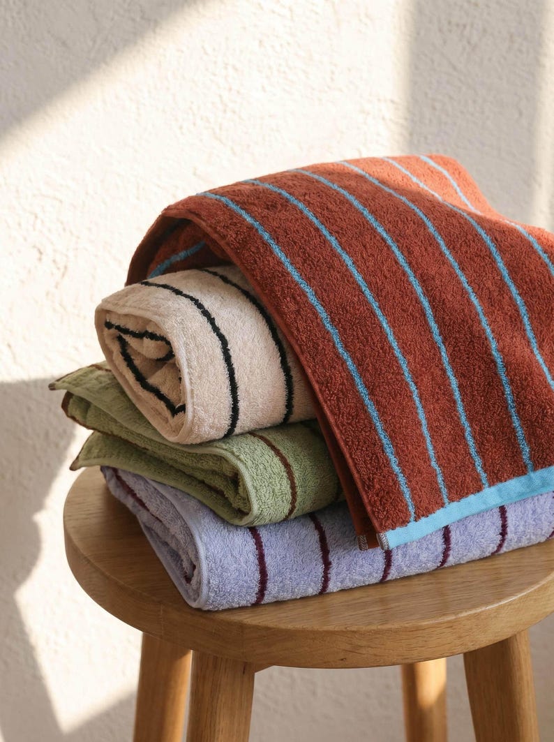 Op de afbeelding: Een stapel van vier gevouwen katoenen handdoeken met verticale strepen. De bovenste handdoek is roestoranje met lichtblauwe strepen, gevolgd door een cr&egrave;mekleurige handdoek met zwarte strepen, een saliegroene met bruine strepen en een lavendelkleurige met donkerpaarse strepen.