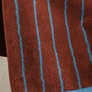 Op de afbeelding: Een textuurrijke, roestkleurige handdoek met verticale lichtblauwe strepen. De handdoek heeft een effen lichtblauwe zoom aan de onderkant en een klein wit waslabel aan de onderste naad, waarop tekst en symbolen zichtbaar zijn.