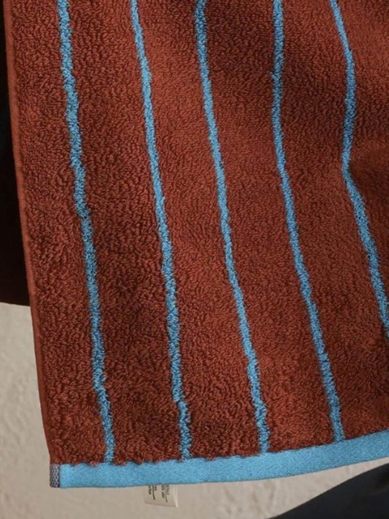 Op de afbeelding: Een textuurrijke, roestkleurige handdoek met verticale lichtblauwe strepen. De handdoek heeft een effen lichtblauwe zoom aan de onderkant en een klein wit waslabel aan de onderste naad, waarop tekst en symbolen zichtbaar zijn.