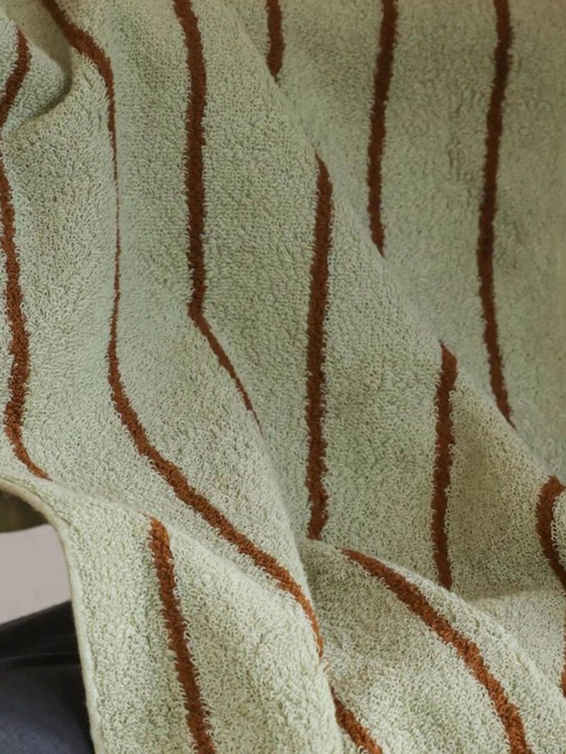 Op de afbeelding: Een zachte, getextureerde handdoek met een licht saliegroene basiskleur en verticale strepen in een diepe roestbruine tint. De stof heeft een luspoolconstructie, wat zorgt voor een pluche, absorberend oppervlak met duidelijke, lineaire patronen.