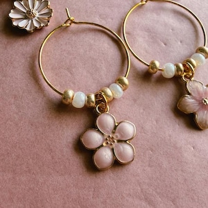 May include: Gold hoop earrings featuring dangling flower charms in shades of light pink and white. Each hoop is accented with small, round, white and gold-toned beads arranged along the bottom curve, creating a delicate and floral jewelry set.